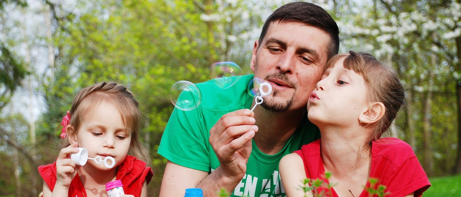 Das Bild zeigt einen Vater mit seinen beiden Töchtern auf einer Blumenwiese beim Seifenblasen spielen.